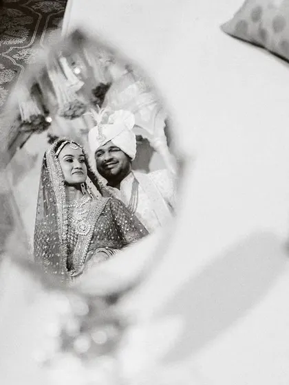 An artistic black and white shot capturing the couple's reflection in a hand mirror, a creative way to frame a classic wedding portrait.