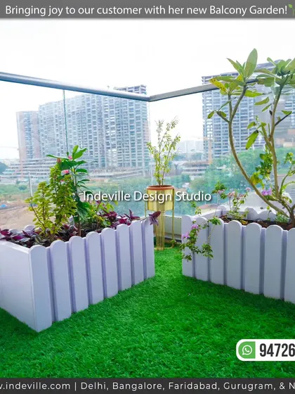 A high-angle view of the custom white planters on a client's balcony. The artificial grass provides a soft, green carpet, and the planters are filled with a mix of flowering plants and small shrubs.