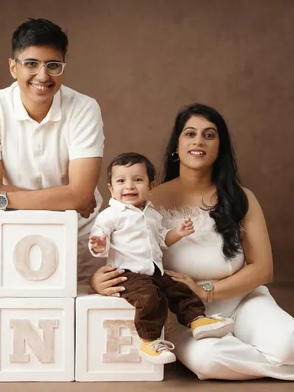A warm family portrait with a brown backdrop, showing two mothers and their son celebrating his first birthday with 'ONE' blocks.