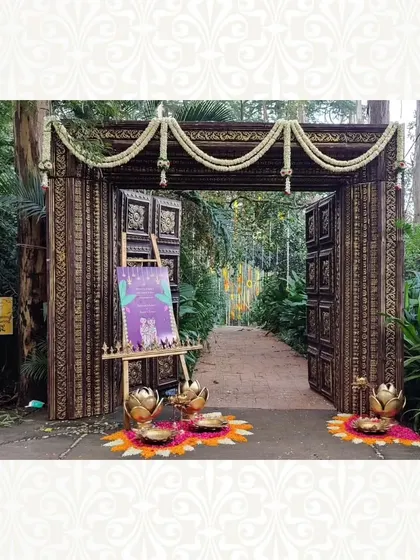 A rustic and traditional wooden gate entrance, decorated with jasmine garlands and brass lotus lamps. This style is perfect for an event with a heritage or temple theme.