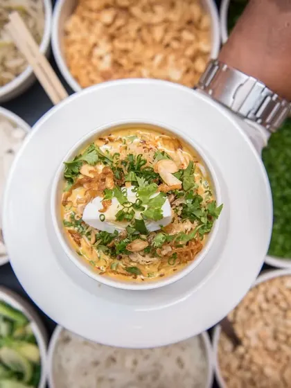 A beautifully garnished bowl of Khow Suey, surrounded by an array of fresh toppings.