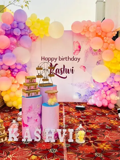 An outdoor butterfly-themed birthday party for Kashvi, with a large backdrop and a rainbow-colored balloon arch.