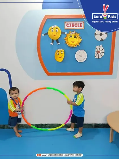 Two young learners work together, holding a large, colorful hula hoop. This 'Circle Day' activity helps them understand shapes and spatial awareness while practicing cooperation and communication.