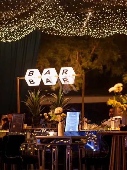 A modern bar sign sits atop a sleek bar, decorated with white florals and set under a canopy of fairy lights. This setup perfectly blends contemporary design with romantic lighting.