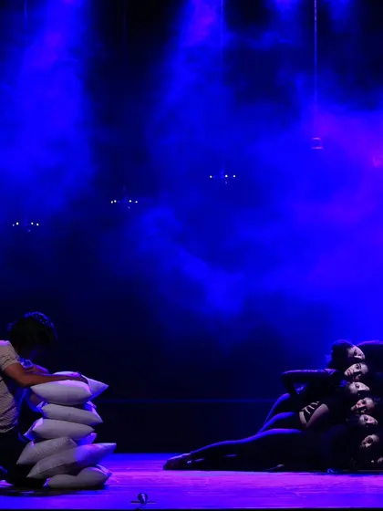 A solo performer stacks pillows in a moment of quiet contemplation from "ME & MY PILLOW." The dramatic lighting focuses attention on the dancer's interaction with the props, telling a story without words.
