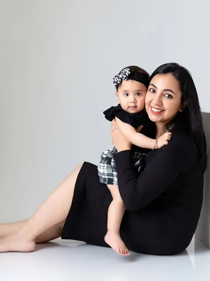 A mother sits on the floor holding her baby girl, both smiling at the camera in a simple, clean studio setting.