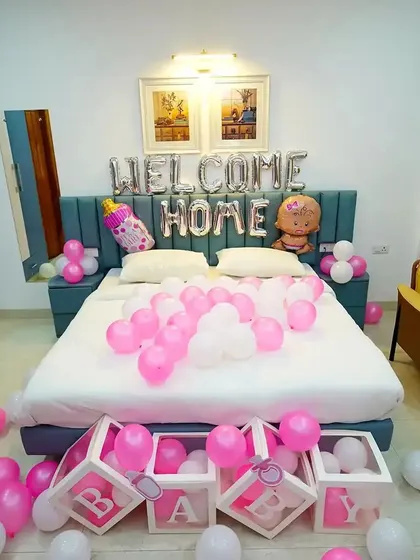 A cozy bedroom setup to welcome the baby home. This decoration includes "WELCOME HOME" foil balloons over the bed, "BABY" blocks filled with pink balloons, and themed foil balloons for a baby girl.