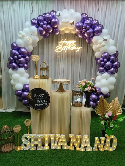 A lovely purple and white themed birthday decoration for Shivanand. The balloon ring is accented with butterfly details, and the setup includes a neon sign and the name in light-up letters on an artificial grass floor.