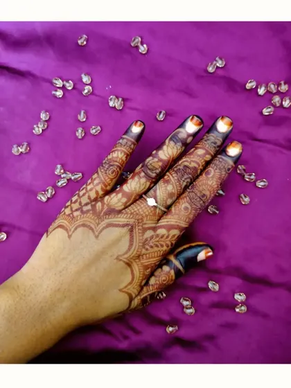 This detailed finger and back-of-hand design shows a scalloped edge at the wrist. The photo is taken against a purple background with crystals, highlighting the rich color of the henna stain.