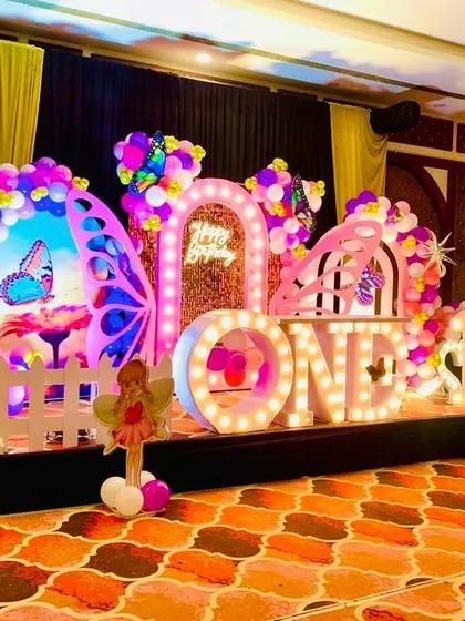 A wide view of a grand butterfly-themed first birthday party in a banquet hall. The stage is decorated with large "ONE" and name marquee letters, a light-up Ferris wheel, and intricate butterfly props.