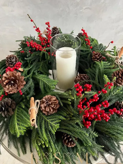 The charming Christmas wreath, perfect for the holidays. This design features fresh pine, bright red ilex berries, natural pinecones, and cinnamon sticks, with a candle in the center for a traditional look.