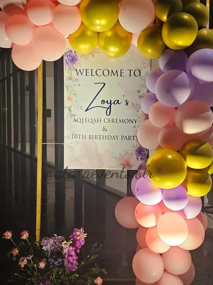A welcome sign for Zoya's Aqeeqah ceremony and 10th birthday party. The sign is framed by a beautiful arch of pink, purple, and gold balloons, creating a festive and celebratory entrance.