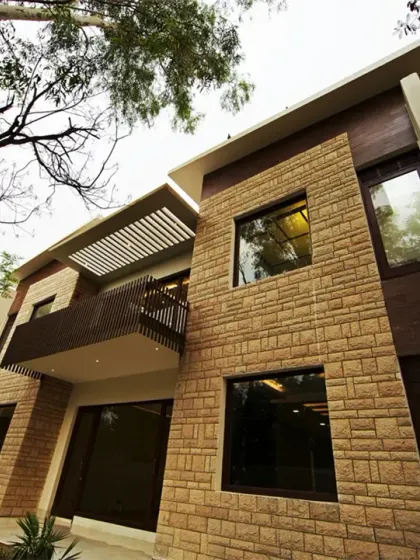 The side elevation of the Sachdeva Farmhouse showcases a modern design with stone cladding and wooden textures. Projecting balconies with wooden louvers and pergola elements add depth, shade, and a rhythmic quality to the façade.