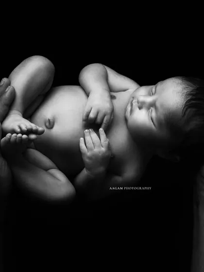 A dramatic and artistic black and white portrait of a newborn resting in their father's hands against a black background. The lighting beautifully sculpts the baby's form.