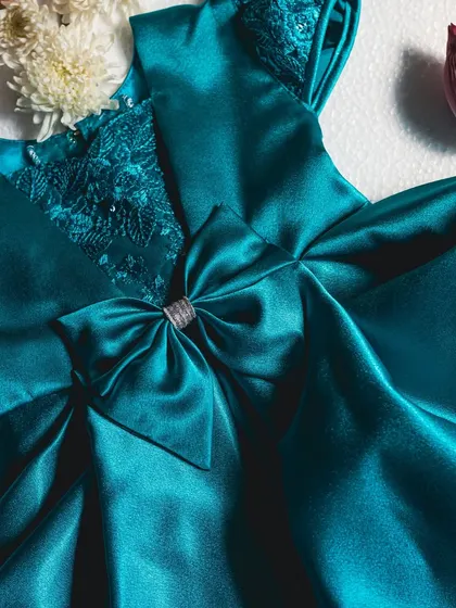 A stunning teal satin dress with intricate lace detailing on the neckline. The large, perfectly constructed bow is the centerpiece of this elegant design.