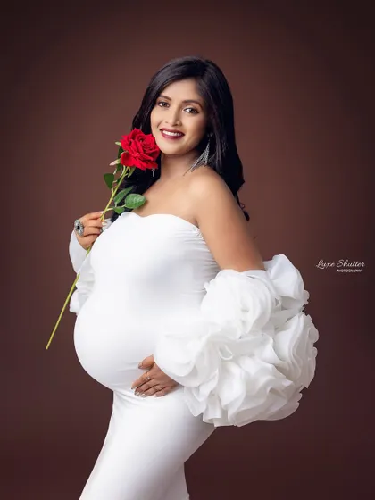A smiling mom-to-be looking radiant in a white gown with statement sleeves, holding a single red rose.