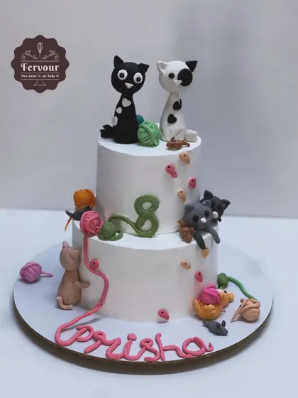 A playful two-tier cat-themed cake for an 8th birthday. The design features cute fondant cats playing with balls of yarn, climbing up the sides of the white whipped cream cake.