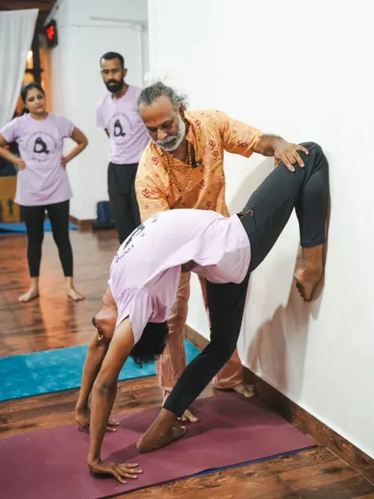 I am providing a hands-on adjustment to a student in a wall-assisted backbend, helping them find their fullest expression of the pose safely.