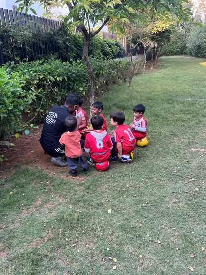 A group of toddlers sits on their footballs, listening intently to their coach under the shade of a tree.