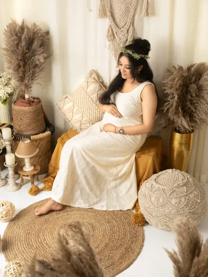 A quiet, contemplative solo portrait. The mother-to-be is seated, looking down at her bump with a gentle smile, surrounded by the soft textures of our boho-chic studio setup.