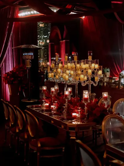 A long shot of the dining tables, featuring towering candelabras and lush red floral arrangements. The mirrored tabletops reflect the candlelight, amplifying the warm and opulent feel of the room.