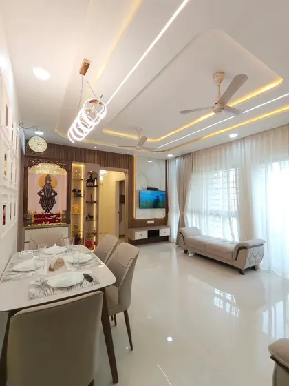An overview of an open-plan living and dining area, showcasing how we create distinct zones within a single space. The dining set is positioned under a modern chandelier, while the living area is defined by a comfortable sofa and a dedicated TV and mandir wall. The glossy floor tiles and consistent lighting scheme tie the entire area together.