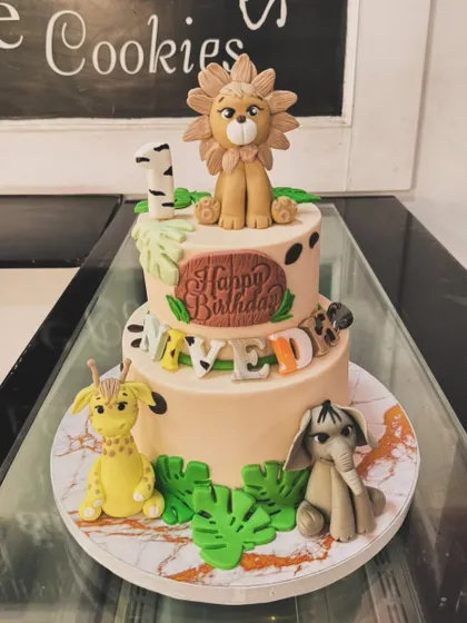 A two-tiered jungle animal cake for a first birthday. This design features a lion, giraffe, and elephant, with a fun mix of animal prints and tropical leaves.