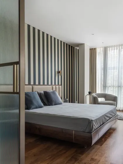 This bedroom is a study in refined comfort, featuring a bold striped wallpaper that adds graphic interest without overwhelming the space. A fluted glass partition provides a subtle separation, enhancing privacy while allowing light to filter through, creating a serene and layered retreat.