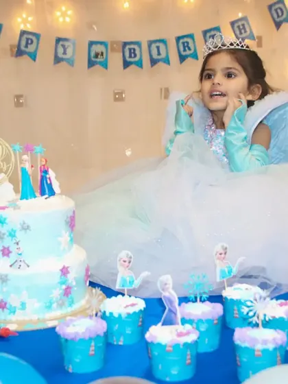 The birthday girl, dressed as a princess, looks on with excitement at her Frozen-themed cake and cupcakes.
