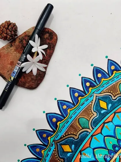 A sneak peek of the 'Peacock in the Desert Rain' mandala, showing a corner of the finished piece with a pen and some natural elements.