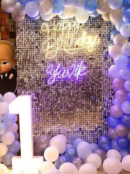 A vibrant Boss Baby themed first birthday party. The decor features a silver shimmer wall, a blue and white balloon arch, and a 'Happy Birthday' neon sign.