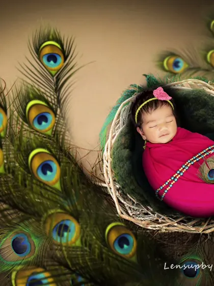 A serene newborn, dressed as little Radha, sleeps peacefully in a nest-like basket adorned with peacock feathers, embodying grace and divinity.