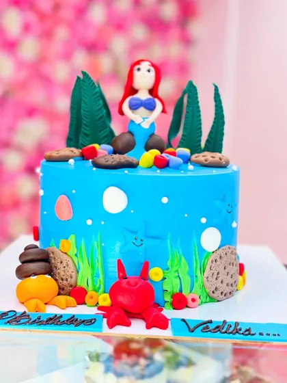 A fun mermaid cake featuring a hand-sculpted Ariel, a friendly crab, and underwater plants. The use of cookie crumbs as 'sand' adds a creative touch.