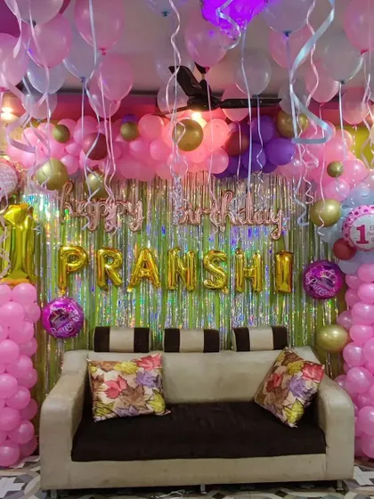 A first birthday party with a full pink and gold balloon setup. The decoration includes a dense balloon arch over the sofa, a shimmer curtain, and themed foil balloons on pillars.