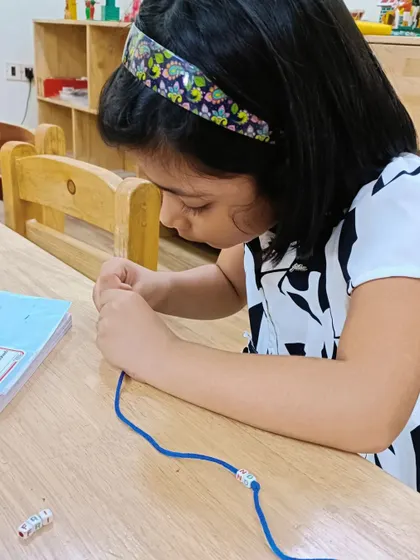 A young girl concentrates on threading beads for her friendship bracelet. This activity requires focus and helps develop hand-eye coordination.