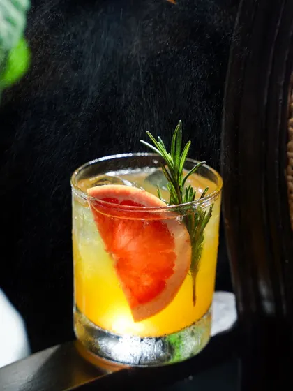 A citrus and rosemary cocktail being garnished with a spritz of zest. The dramatic lighting from behind illuminates the spray, adding a sense of action and freshness to this beverage photograph.