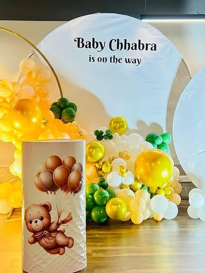 A teddy bear-themed baby shower with a "Baby Chhabra is on the way" message. The decor includes a large moon prop and a balloon arch in yellow, green, and white.