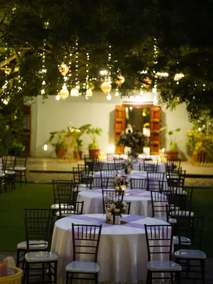 An enchanting garden dining setup. Tables are arranged under a canopy of trees adorned with hanging lanterns and fairy lights, creating a magical and intimate atmosphere for an outdoor reception.