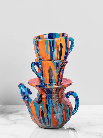 This playful take on a teapot sculpture features a stack of cups and a pot with a bird-shaped spout, all finished in a vibrant blue, orange, and red drip-paint style. It's a cheerful accent for your kitchen or dining area.