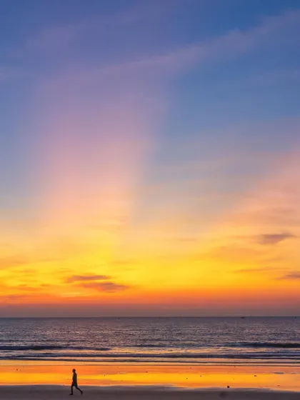 A lone walker on the beach during a spectacular winter sunset. The sky is filled with vibrant yellows, oranges, and purples, creating a breathtaking scene.