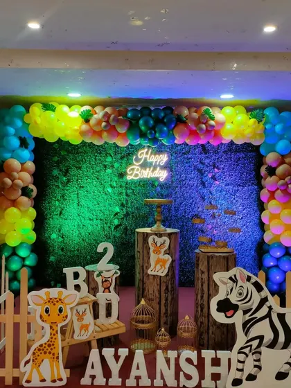 A colorful jungle party setup with a vibrant, multi-colored balloon garland over a grass backdrop. The scene is lit with fun colors and features cute animal props.