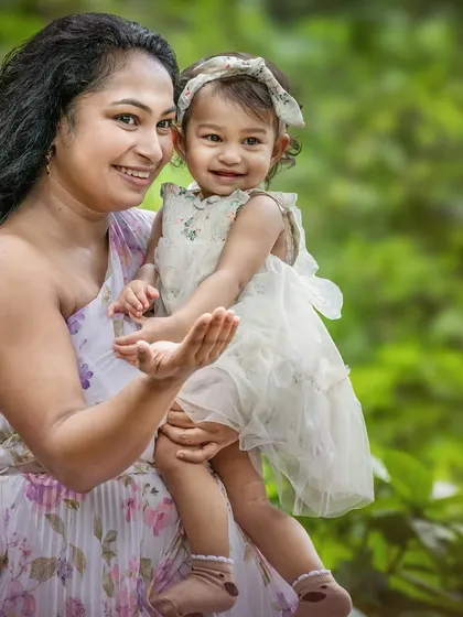 A mother holds her young daughter, both smiling warmly at the camera against a vibrant green, natural background. This is a classic, joyful mother-daughter portrait that feels both candid and timeless.