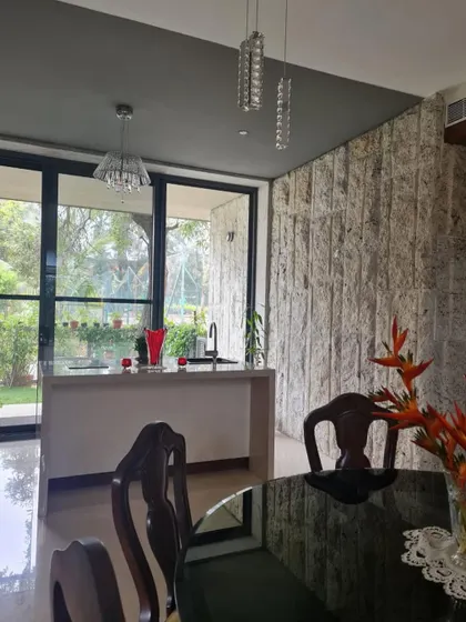 The dining area in "Casa Magnifera" is defined by a rough, natural stone wall and a full-length glass window looking out onto the garden, bringing texture and nature to the table.