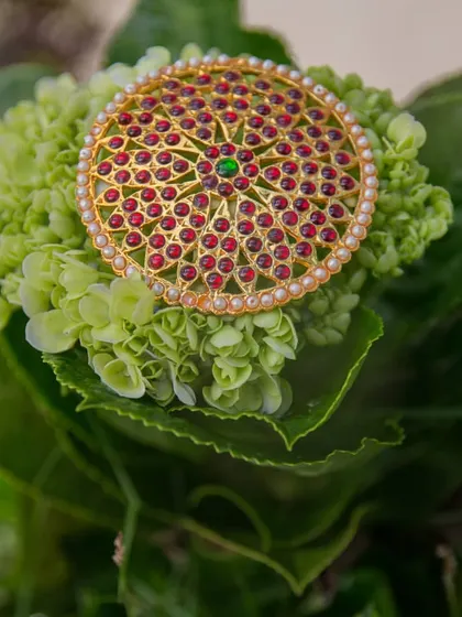 A detailed photograph of a 'jadai billai', a traditional hair ornament used in South Indian weddings. We focus on capturing the intricate red and green stonework and pearl details.