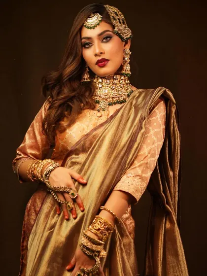 A full portrait of a royal bride in a golden-bronze saree. The look is accessorized with heavy green and gold jewellery, showcasing a rich and opulent aesthetic.