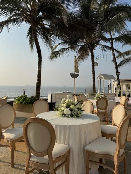 Elegant guest seating by the sea, with simple white tablecloths and rustic wooden chairs. A classic white floral centerpiece completes the look of understated coastal elegance.