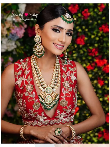 A smiling bride against a floral backdrop, looking radiant in her red lehenga and statement kundan jewellery set.