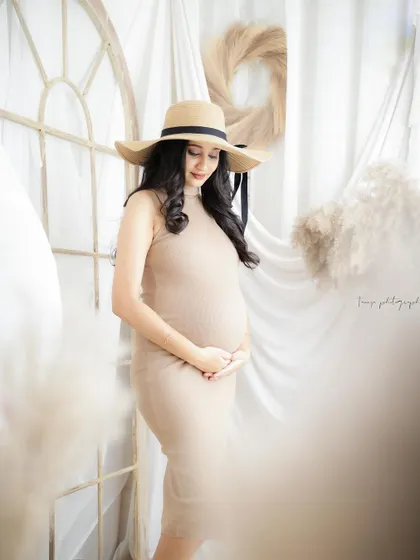 A chic and serene portrait. The mom-to-be wears a simple beige dress and a sun hat, creating a timeless and elegant look.