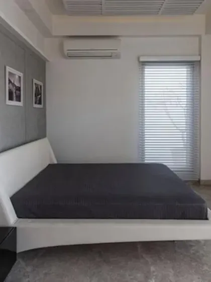 This bedroom in J House has a minimalist, modern design. A low white platform bed, grey textured accent wall, and large windows with blinds keep the space uncluttered and filled with natural light.