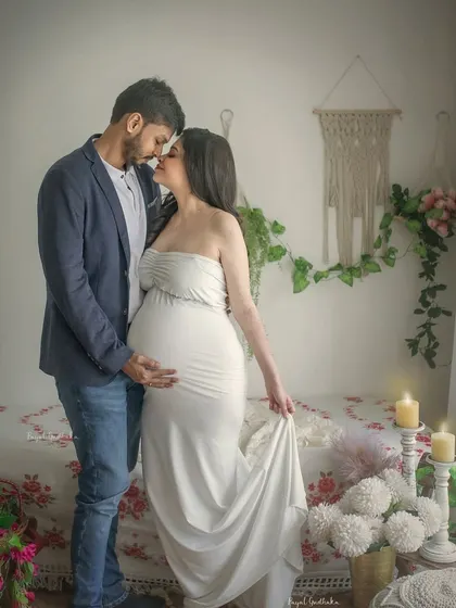 An intimate portrait of an expecting couple in a boho-styled studio setting. The soft natural light from the window and their close embrace create a romantic and dreamy atmosphere.